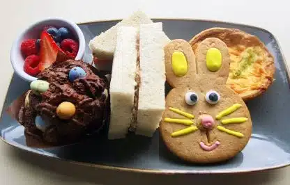 Children's Afternoon Tea on a blue plate. Small pot of fresh fruit, chocolate krispy cake with mini eggs, ham sandwich cut in rectangles, cheese quiche & biscuit in shape of a rabbit face with icing to portray the rabbit face.